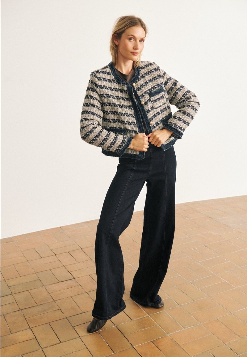 Femme portant une veste rayée texturée avec de gros boutons et un pantalon noir à jambes larges, debout sur un sol carrelé contre un mur blanc.