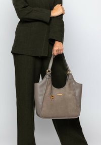Sac à main en cuir et daim gris de forme arrondie, avec quincaillerie dorée et un accent porte-clés. Tenue par une personne vêtue de vêtements vert olive.