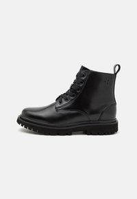 Botas de tobillo de cuero negro con diseño de cordones, puntera redonda y suela de goma resistente. Presenta un detalle del logotipo en el lado exterior.