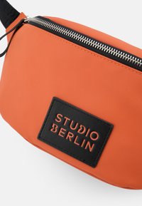Orange Nylon-Bauchtasche mit strukturiertem Oberflächen, ausgestattet mit einem schwarzen Logo-Patch und einem silbernen Reißverschluss entlang der oberen Kante.