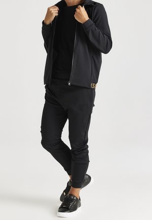 Mann trägt schwarze Jacke mit Reißverschluss, schwarze Hose und schwarze Sneaker mit weißen Sohlen auf weißem Hintergrund.