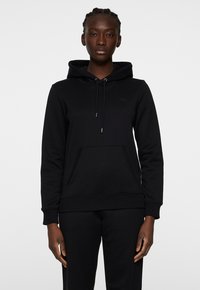 Svart huvtröja i mjukt material, med en framficka, ribbade ärmslut och en subtil logga på bröstet. Enkelt design.