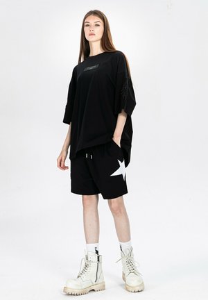 Joven mujer con camiseta negra extragrande, pantalones cortos negros con estrella blanca, calcetines blancos y botas blancas con cordones, de pie frente a un fondo liso.