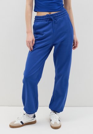 Joggers de color azul royal con cintura elástica y cordón. Puños acanalados en los tobillos. Combinados con zapatillas blancas. Textura de tela suave.