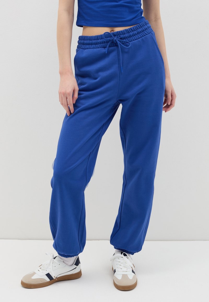 Koningsblauwe joggers met een elastische tailleband en trekkoord. Geribbelde manchetten bij de enkels. Gepaard met witte sneakers. Gladde stoftextuur.