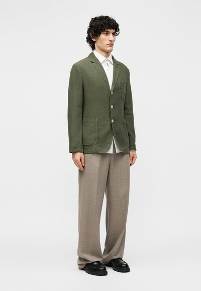 Άνδρας που στέκεται φορώντας ένα χακί πράσινο σακάκι, λευκό πουκάμισο, μπεζ φαρδιά παντελόνια και μαύρα παπούτσια μπροστά σε ένα απλό φωτεινό φόντο.