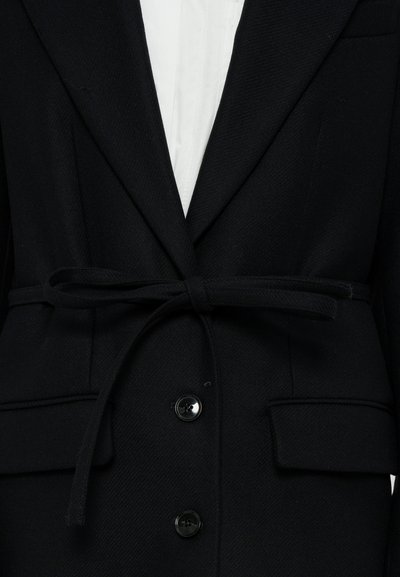 Blazer preta com tecido texturizado, apresentando um cinto atado na cintura, lapelas com entalhe e dois botões frontais. Camisa branca visível por baixo.