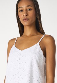 White cotton camisole with thin straps, featuring embroidered eyelet patterns and a loose fit. Smooth texture, V-neckline, and lightweight design.