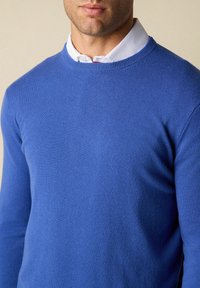 Modrý svetr s crew-neck výstřihem vyrobený z měkkého materiálu, s hladkým povrchem a dlouhými rukávy. Nesený přes bílou košilovou košili.