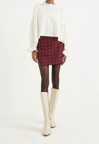 Top bianco a maniche lunghe con rouches, abbinato a una mini gonna a scacchi rossi. La modella indossa stivali alti fino al ginocchio, crema, con zip e tacco. Texture lisce e motivi distintivi.