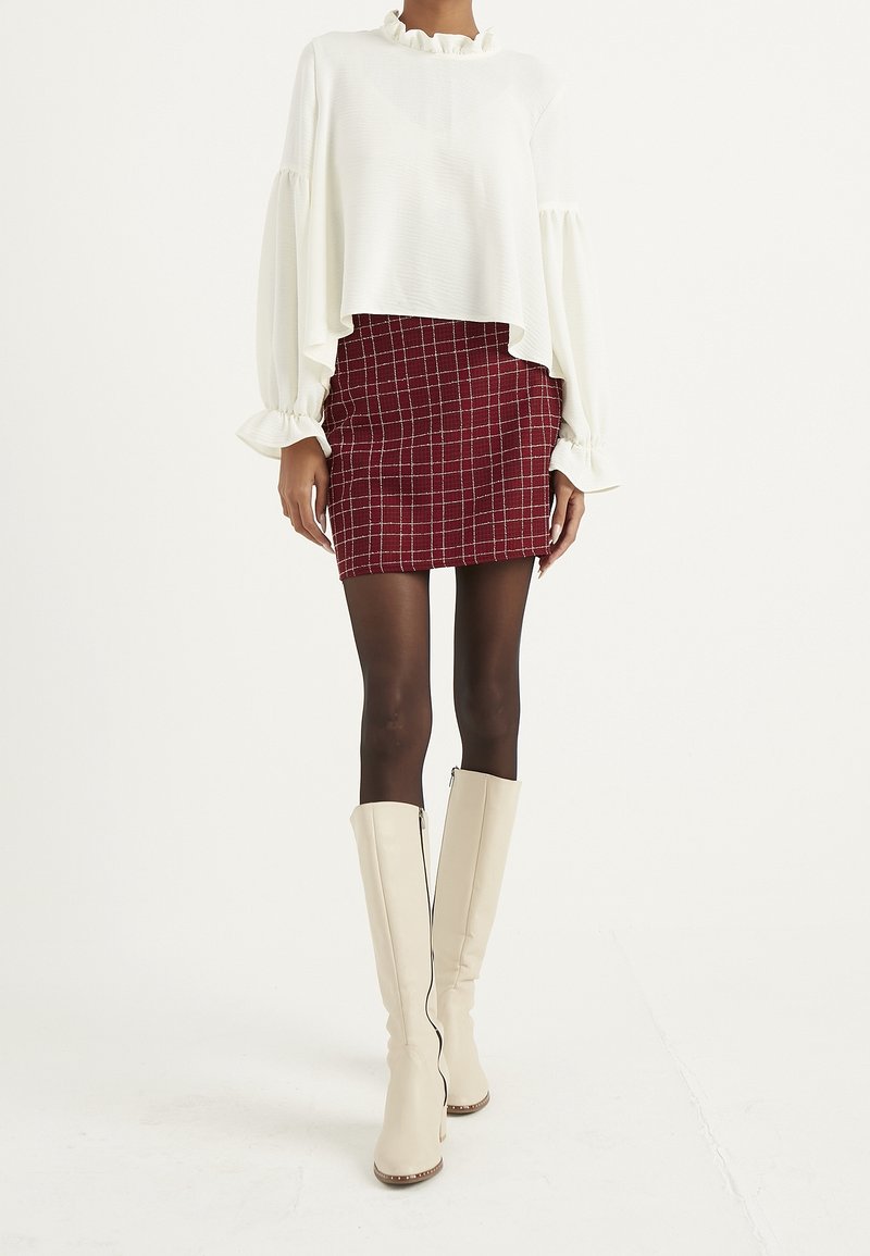Top bianco a maniche lunghe con rouches, abbinato a una mini gonna a scacchi rossi. La modella indossa stivali alti fino al ginocchio, crema, con zip e tacco. Texture lisce e motivi distintivi.