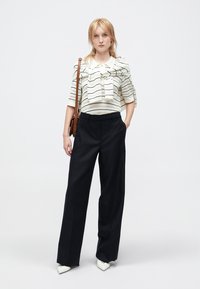 Valkoinen ja musta raidallinen paita, jossa on nauhatyyny, yhdistettynä korkeavyötäröisiin mustiin leveälahkeisiin housuihin. Malli pitää kädessään ruskeaa olkalaukkua.