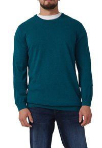Türkis gestrickter Pullover mit Rundhals, langen Ärmeln und gerippten Manschetten. Über einem weißen Hemd getragen, kombiniert mit dunklen Jeans.