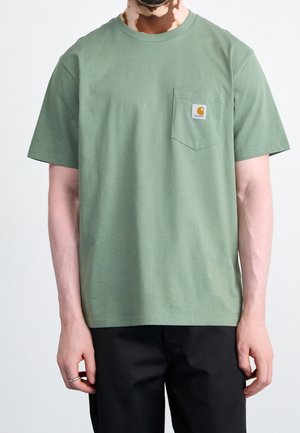 Mężczyzna noszący zieloną koszulkę z krótkim rękawem z kieszenią na piersi z logo Carhartt, w połączeniu z czarnymi spodniami.