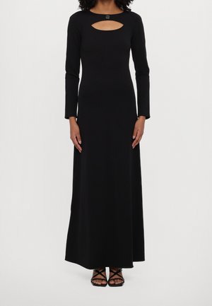 Woman wearing a long black dress with long sleeves and a small oval cutout below the neckline, paired with black strappy sandals.