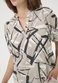 Kvinde iført en beige bluse med mønster i sort og hvidt abstrakt linjemønster og korte puffærmer, hånden hviler i lommen.