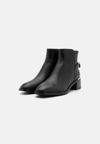 Bottines en cuir noir avec un bout pointu, un talon block bas et une sangle latérale avec des détails en métal argenté. Texture lisse, design élégant.