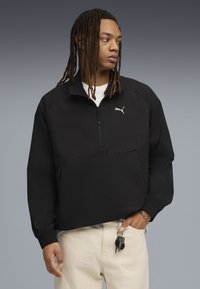 Giacca pullover nera Puma con collo alto zip, corpo testurizzato e polsini elasticizzati. Indossata sopra una camicia bianca con le chiavi visibili.