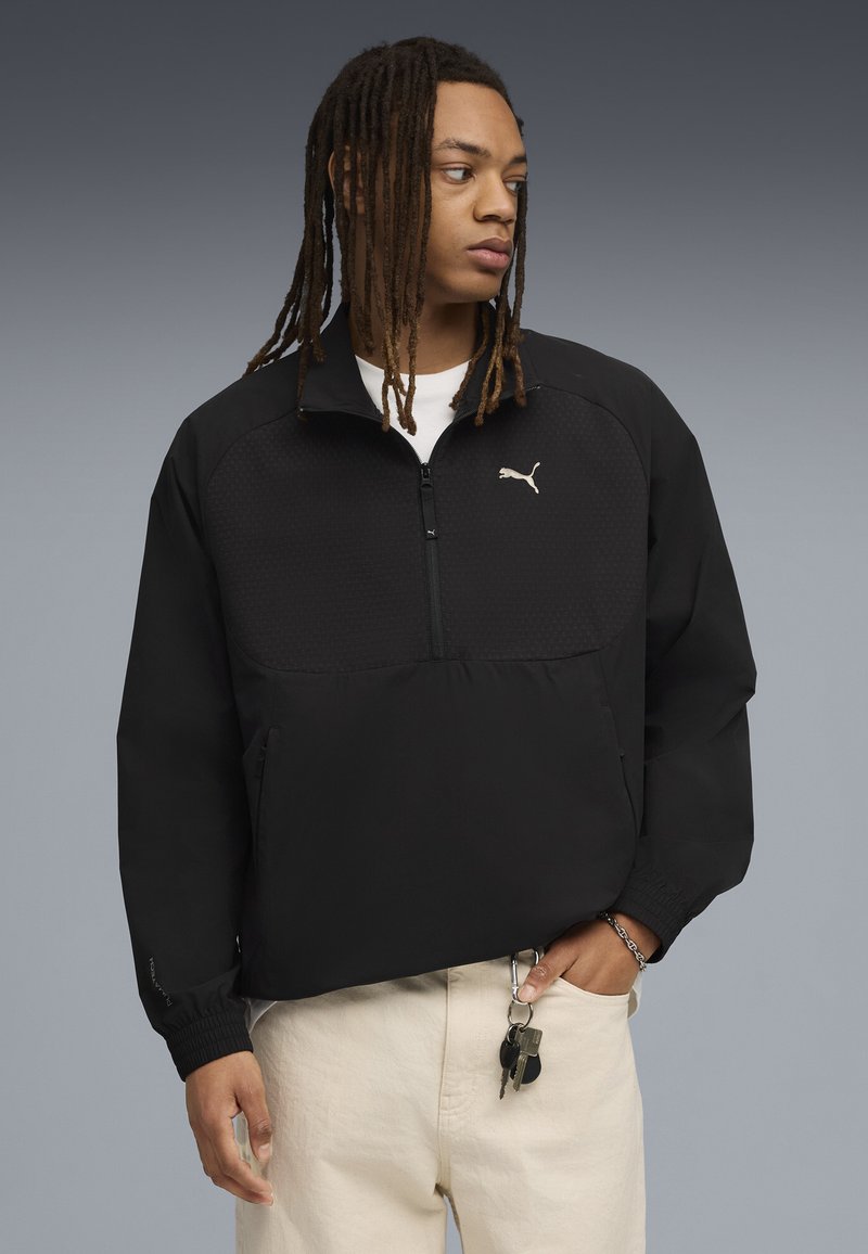 Giacca pullover nera Puma con collo alto zip, corpo testurizzato e polsini elasticizzati. Indossata sopra una camicia bianca con le chiavi visibili.