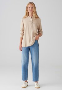 Beige Langarmhemd mit Henley-Ausschnitt und Knöpfen, kombiniert mit hellblauen Röhrenjeans und weißen Sneakers. Glatte Textur.