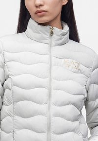 Femme portant une veste matelassée gris clair avec un col haut et un logo EA7 Emporio Armani visible sur la poitrine, fermeture éclair complètement fermée.