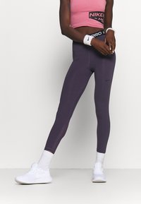 Crop top Nike Pro w jasnoróżowym kolorze z logo, zestawiony z dopasowanymi fioletowymi legginsami z przeszyciami i elastycznym pasem z logo. Białe sneakersy.