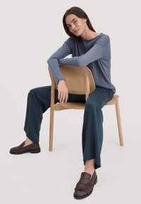 Blauwe lange mouw top, teal wijde broek en bruine franjes loafers. Model zit op een houten stoel, met een zachte studio achtergrond.