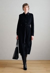 Černé šaty s dlouhým rukávem na knoflíky, se zavázáním v pase a hladkou texturou. Kombinovány s černými kozačkami až ke kolenům a koženou kabelkou přes rameno.