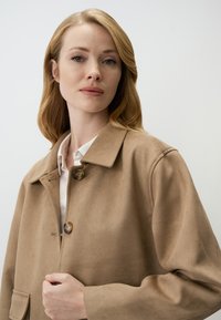 Giacca in suede beige con colletto, chiusura a bottoni e due tasche frontali. Presenta una texture morbida e una vestibilità rilassata.