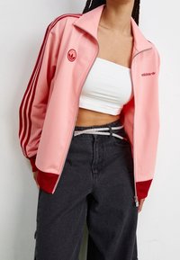 Persona che indossa una giacca a track di Adidas rosa con strisce rosse, una canotta bianca corta, jeans scuri a vita alta e una cintura di strass.