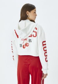 Sudadera blanca recortada con texto rojo, que presenta un gráfico de cereza y un lazo. La tela parece suave y lisa, con capucha y cordón.