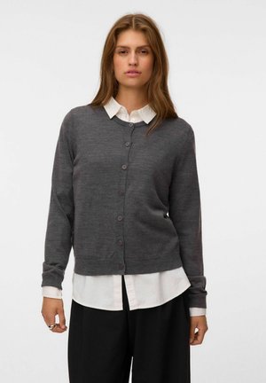 Jeune femme aux longs cheveux bruns portant un cardigan gris foncé boutonné par-dessus une chemise blanche à col et un pantalon noir sur un fond uni.