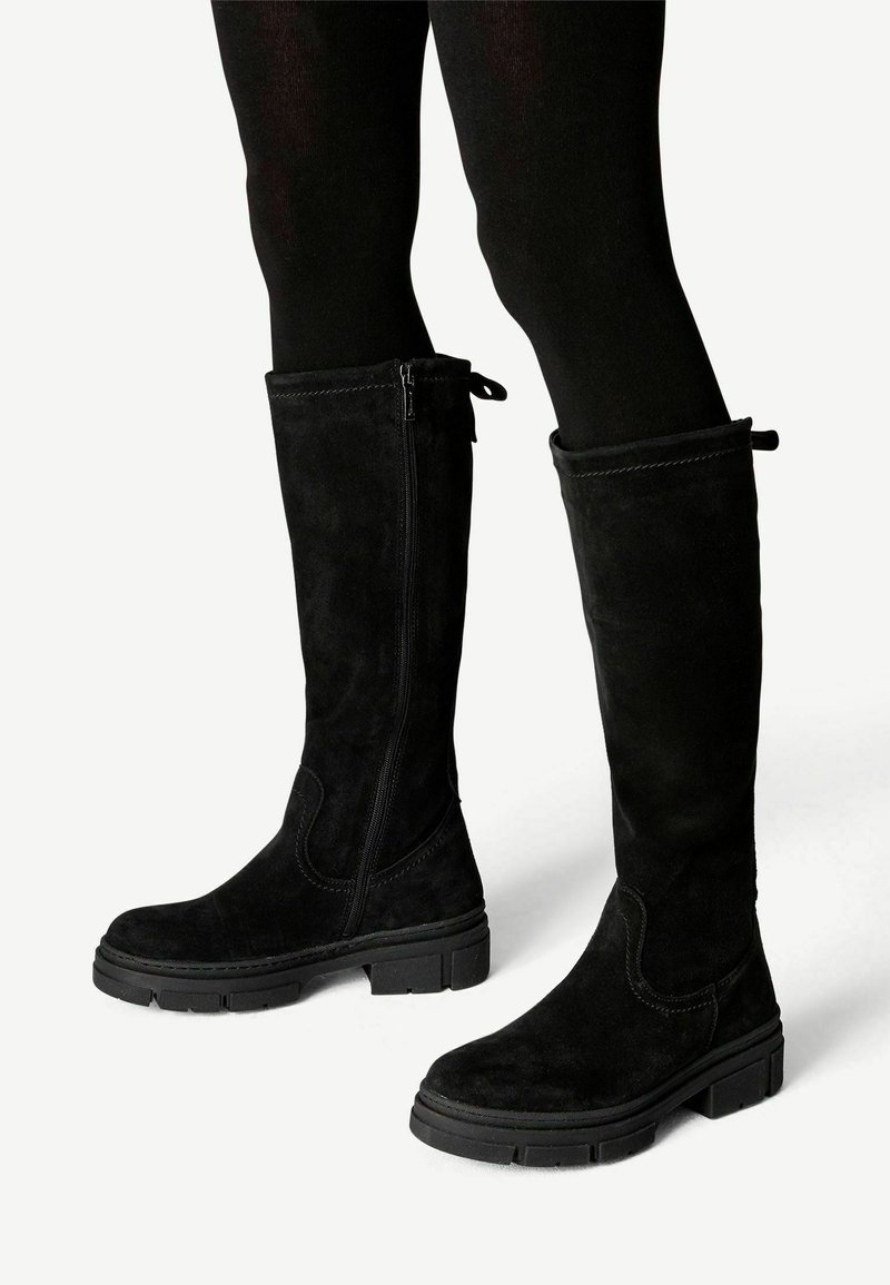 Bottes montantes en daim noir avec fermetures éclair latérales et semelles en caoutchouc épaisses, portées avec des collants noirs sur un fond clair.