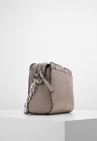 Malá taupe kožená crossbody taška se stříbrným řetízkem a zipem, vystavená na bílém podstavci na pozadí bez vzoru.