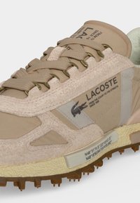 Μπεζ αθλητικό παπούτσι Lacoste με υφή υφάσματος, καφέ κορδόνια, λογότυπο με επωνυμία και κείμενο, και λαστιχένια σόλα με ορατές αντιολισθητικές ακίδες.
