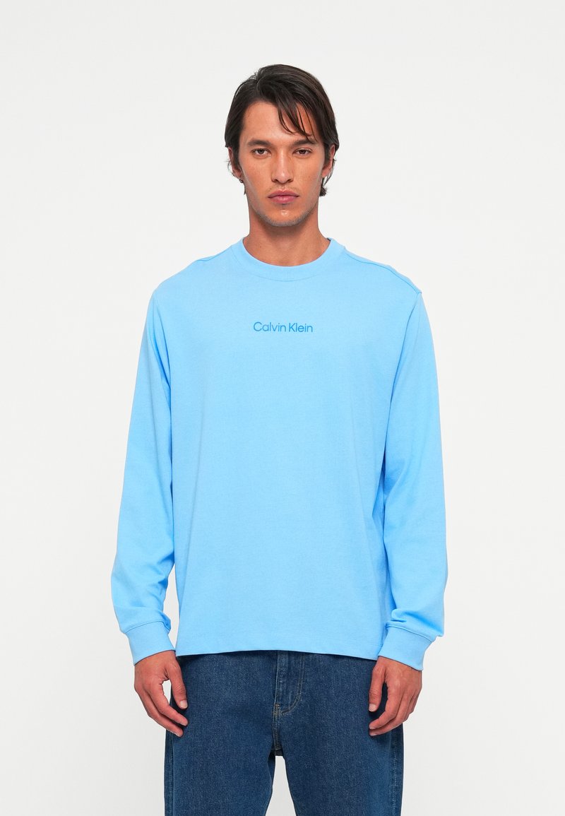 Bărbat îmbrăcat cu o cămașă cu mânecă lungă de culoare bleu deschis, având logo-ul "Calvin Klein", și blugi de culoare bleu închis, stând în fața unui fundal alb simplu.