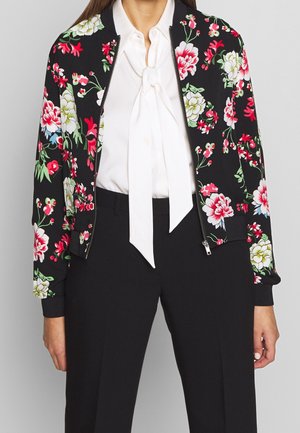 Mujer con cazadora bomber negra con estampado floral y cremallera, sobre blusa blanca con lazo, combinada con pantalones negros hechos a medida.