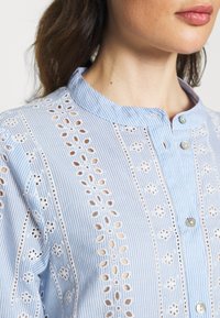 Blusa riscada azul claro com colarinho mandarim, apresentando detalhes de ilhós bordados, pequenos botões e um tecido texturizado.