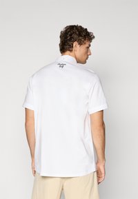 Hvit kortermet polo-shirt med krage, med en liten svart "Balloon Golf" logo på baksiden. Glatt, lett stoff.
