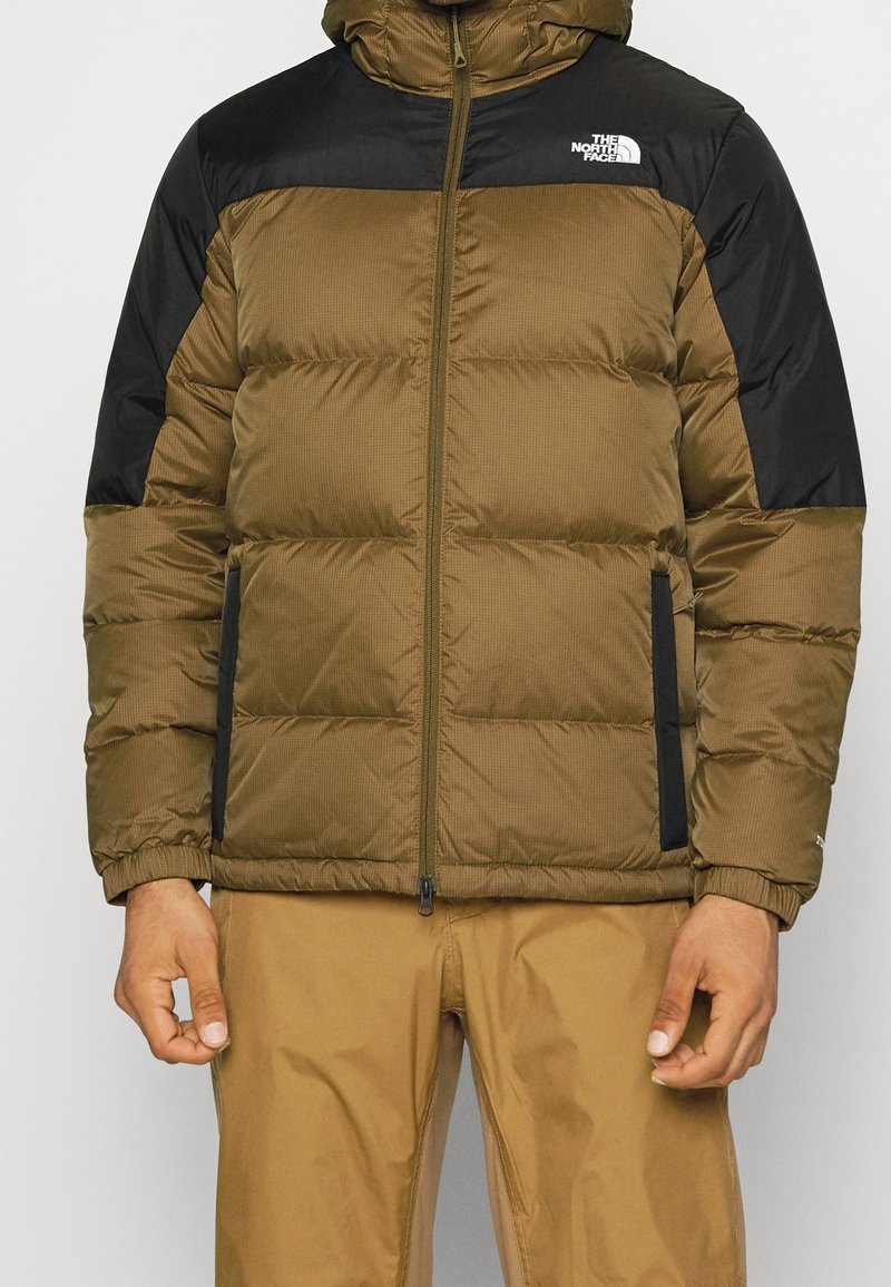 Vadderad jacka i olivgrön med svarta axlar, med en hel dragkedja, två sidofickor och en huva. Texturerad tyg visar ett subtilt mönster.