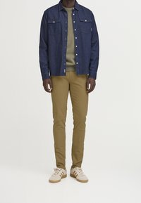 Dunkelblaue Jeanshemd mit zwei Brusttaschen, olivgrüner Pullover, beige schmale Hose und cremefarbene Sneakers mit braunen Akzenten.