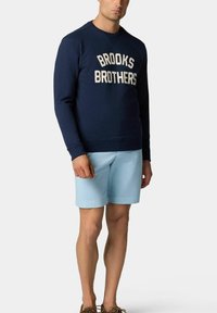 Námorně modrá mikina s logem "BROOKS BROTHERS" v bílé barvě, spárovaná se světle modrými šortkami a hnědými botami, stojící na jednobarevném pozadí.