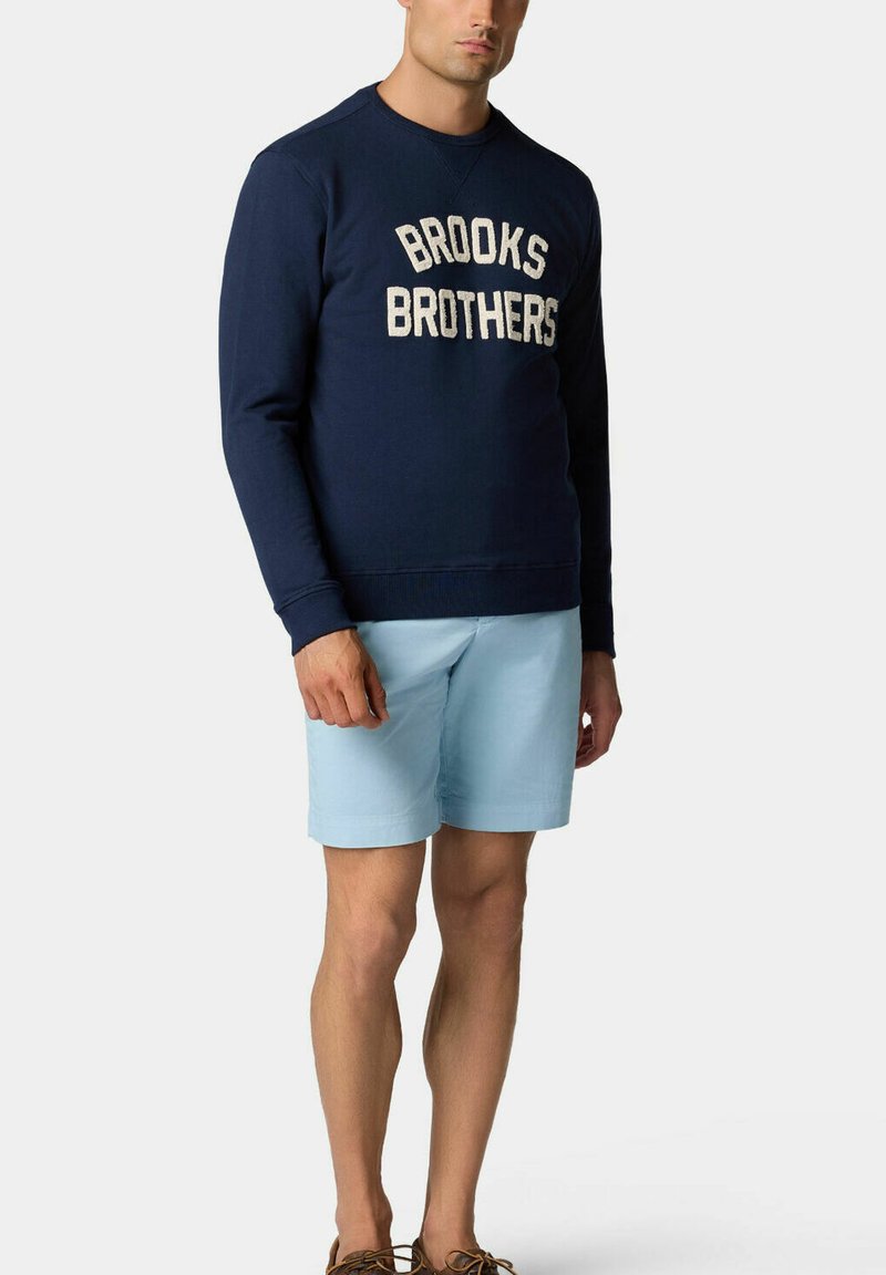 Námorně modrá mikina s logem "BROOKS BROTHERS" v bílé barvě, spárovaná se světle modrými šortkami a hnědými botami, stojící na jednobarevném pozadí.