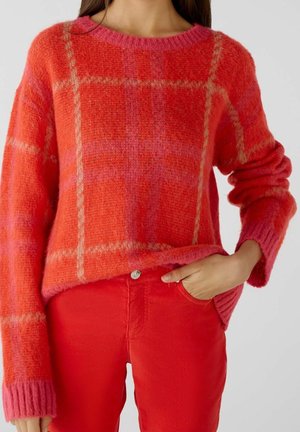 Kvinde iført en blød rød og pink ternet sweater med røde bukser, hånden i lommen, foran en ensfarvet lys baggrund.