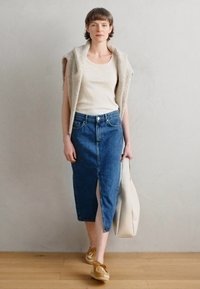 Gilet lavorato a maglia beige sopra una canotta bianca a coste, abbinato a una gonna a tubino in denim blu a lunghezza ginocchio con spacco frontale e borsa tote beige.
