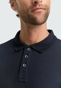 Dunkelblaues, geripptes Poloshirt mit klassischem Kragen, featuring drei Knopfverschluss und kontrastierenden runden Knöpfen. Texturierte Stoffdetails sichtbar.