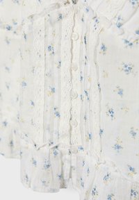 Blouse blanche avec un petit imprimé floral bleu et jaune, bordure en dentelle verticale, bords volantés et boutons blancs sur le devant.