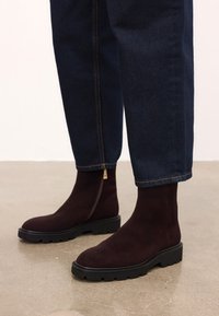 Temno rjave antilop škornje do gležnjev s masivnimi črnimi podplati in stranskimi zadrgami, oblečene z temno modrimi jeans hlačami na gladki tleh.