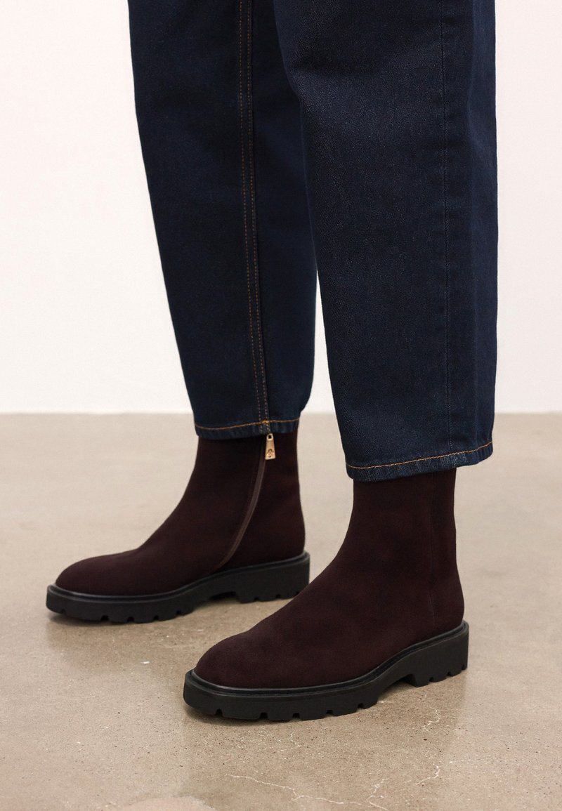 Temno rjave antilop škornje do gležnjev s masivnimi črnimi podplati in stranskimi zadrgami, oblečene z temno modrimi jeans hlačami na gladki tleh.