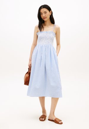 Kobieta ubrana w niebiesko-białą, paskowaną, midi sukienkę bez rękawów z marszczonym gorsetem, trzymająca brązową torebkę i nosząca brązowe sandały na płaskiej podeszwie.